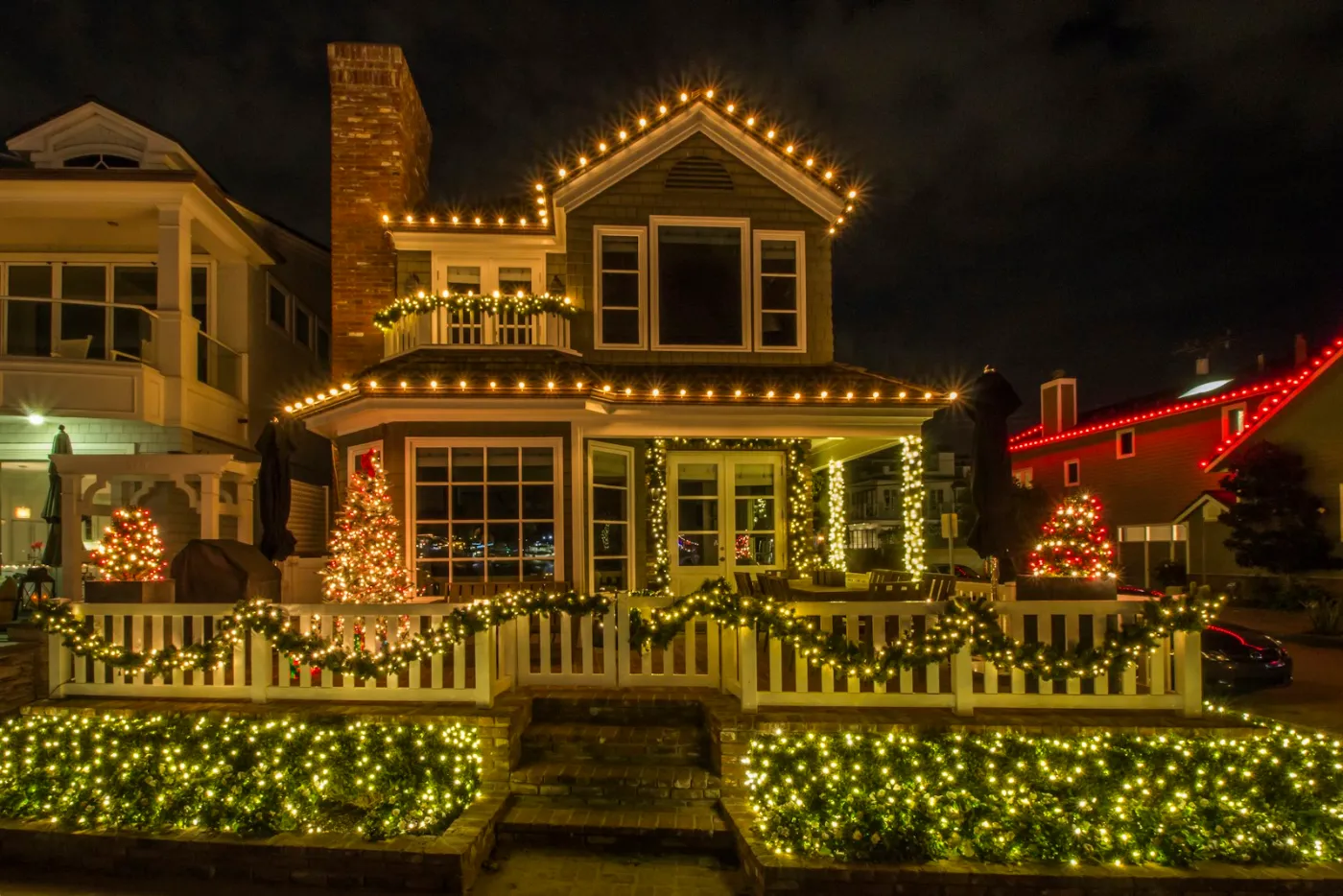 Coastal home decorated with Christmas lights along the roofline, porch, fence, and net-lit shrubs